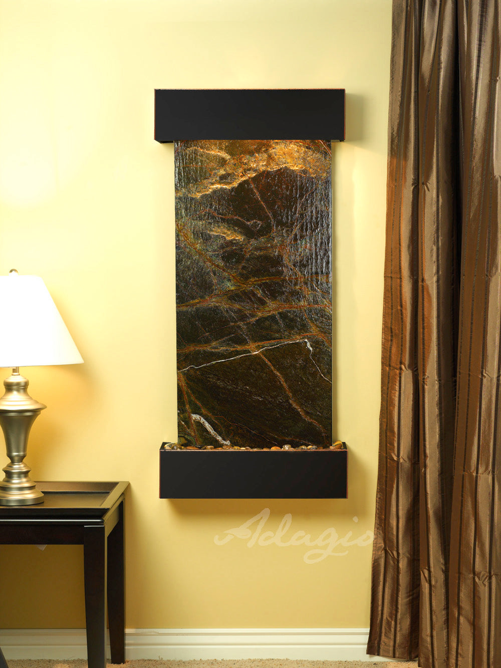 Wall-mounted water feature with green-rainforest marble on a yellow wall, next to a lamp and curtain.