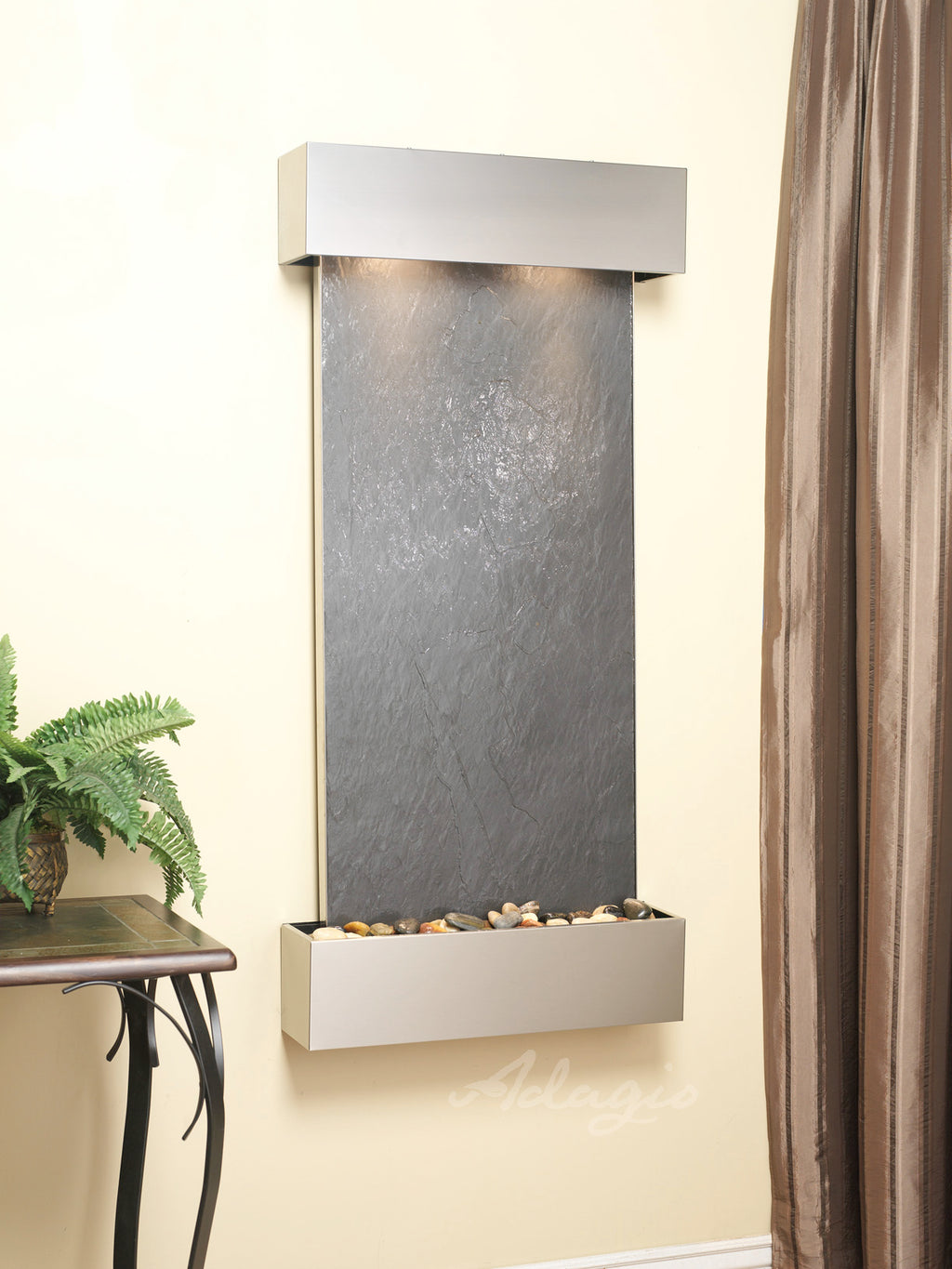 Wall-mounted water feature with dark slate surface and pebbles on a beige wall.
