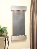 Wall-mounted water feature with dark slate surface and pebbles on a beige wall.