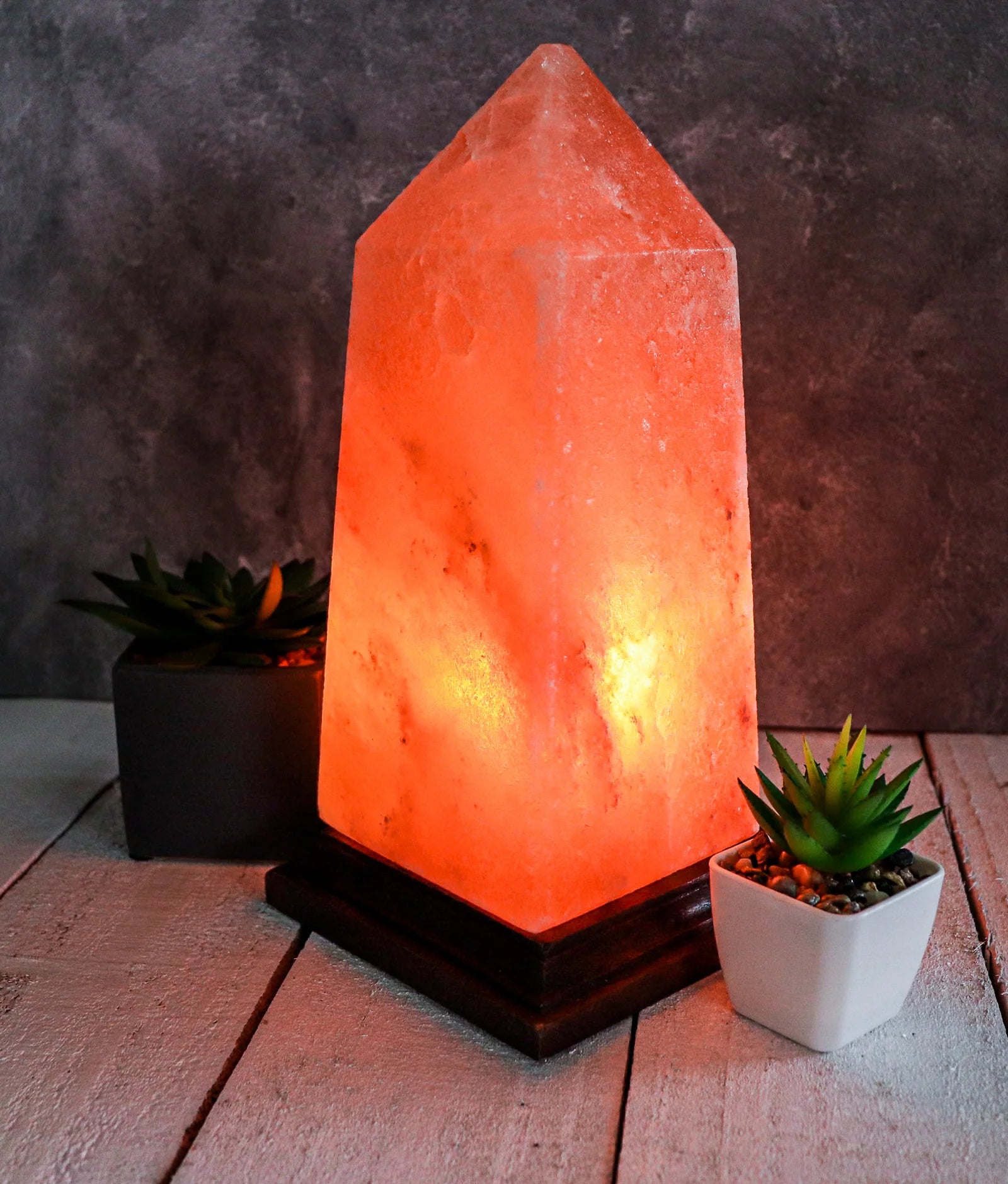 Himalayan salt lamp on a wooden surface with two small plants, against a dark textured wall.